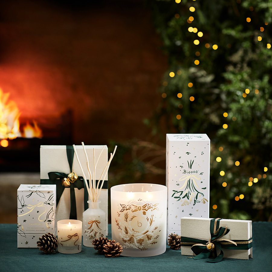 Christmas edition candle sets from Bamford placed on a green table, with a fireplace and lit up Christmas trees in the background. 