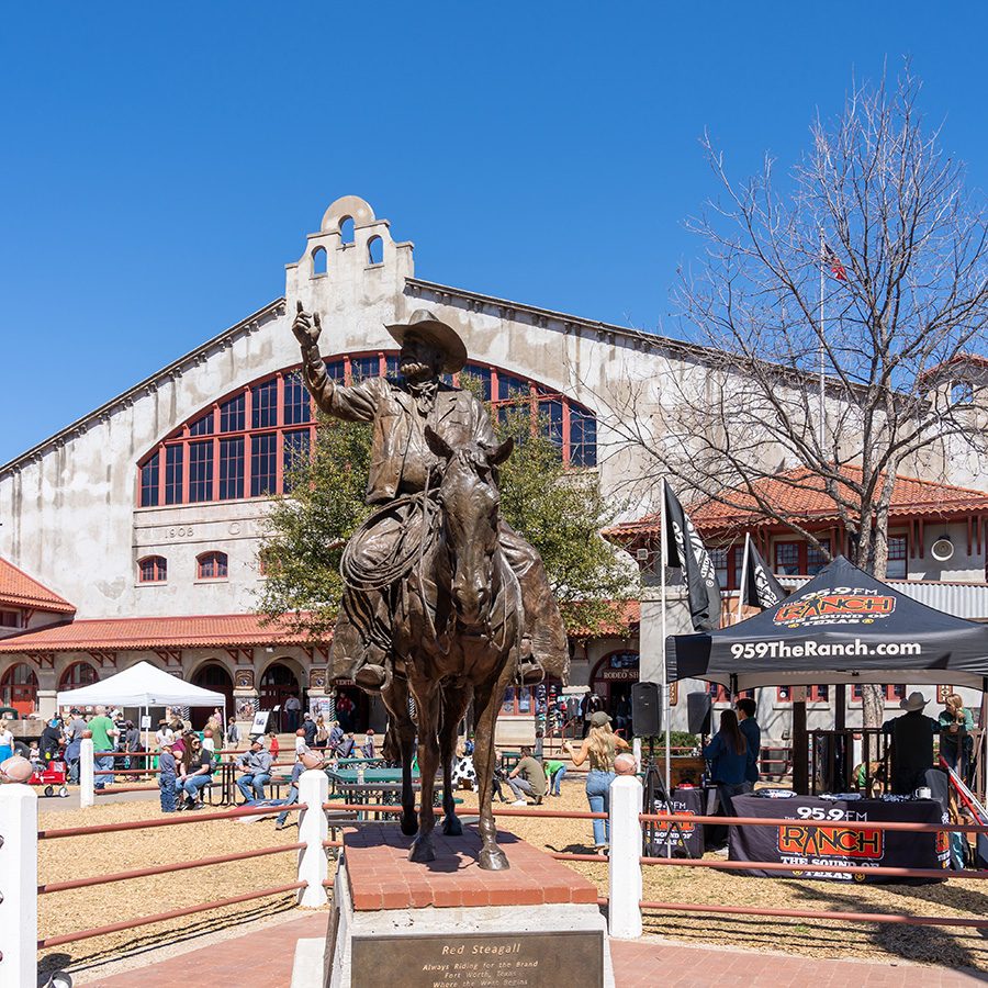 A statue of a man on a horse stands in front of the Cowtown Coliseum, a large grey building with a wide arched window.