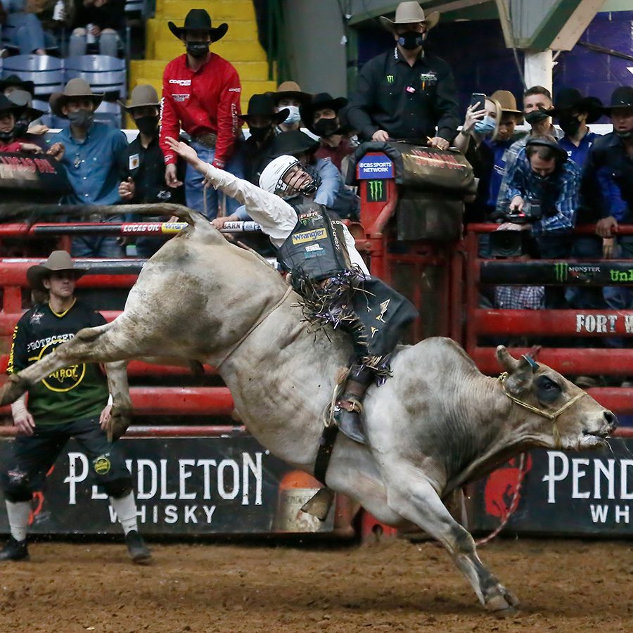 Inside the Cowtown Coliseum, a bull with a person sitting on its back kicks its hind legs, while crowds look on from the stands.