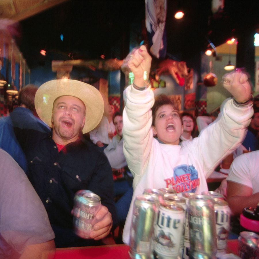 People cheer while standing at a bar inside a crowded dance hall 