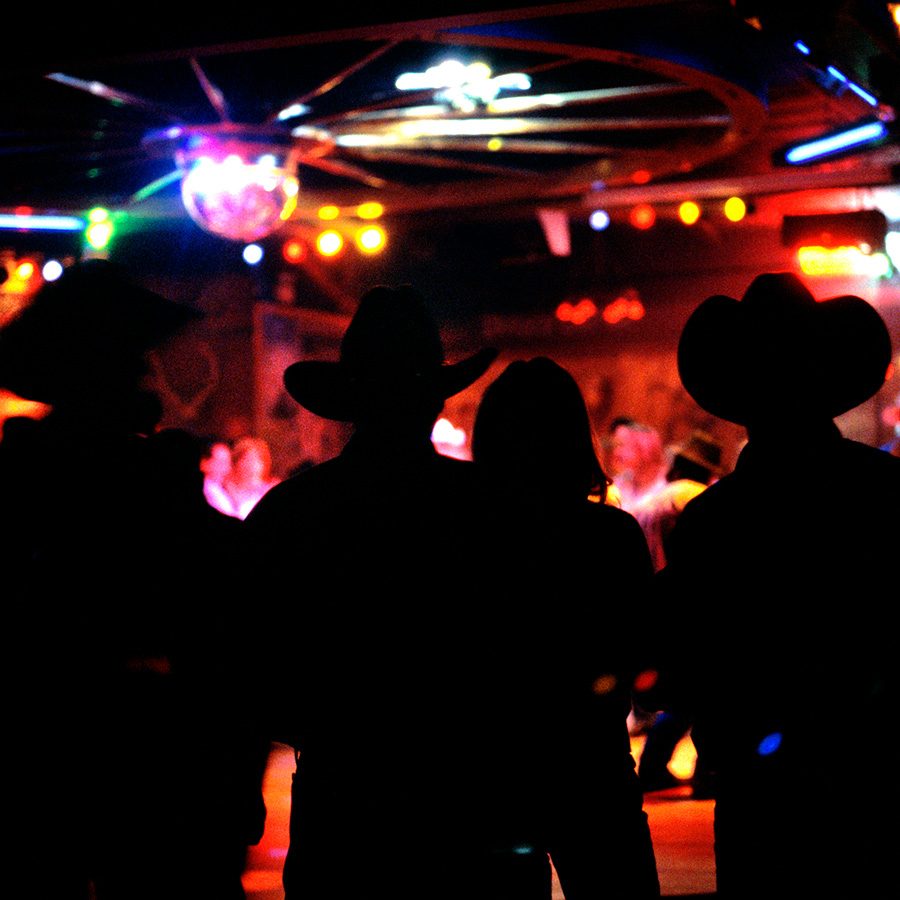 The silhouette of a group of people wearing cowboy hats at a dancehall lit by disco lights     