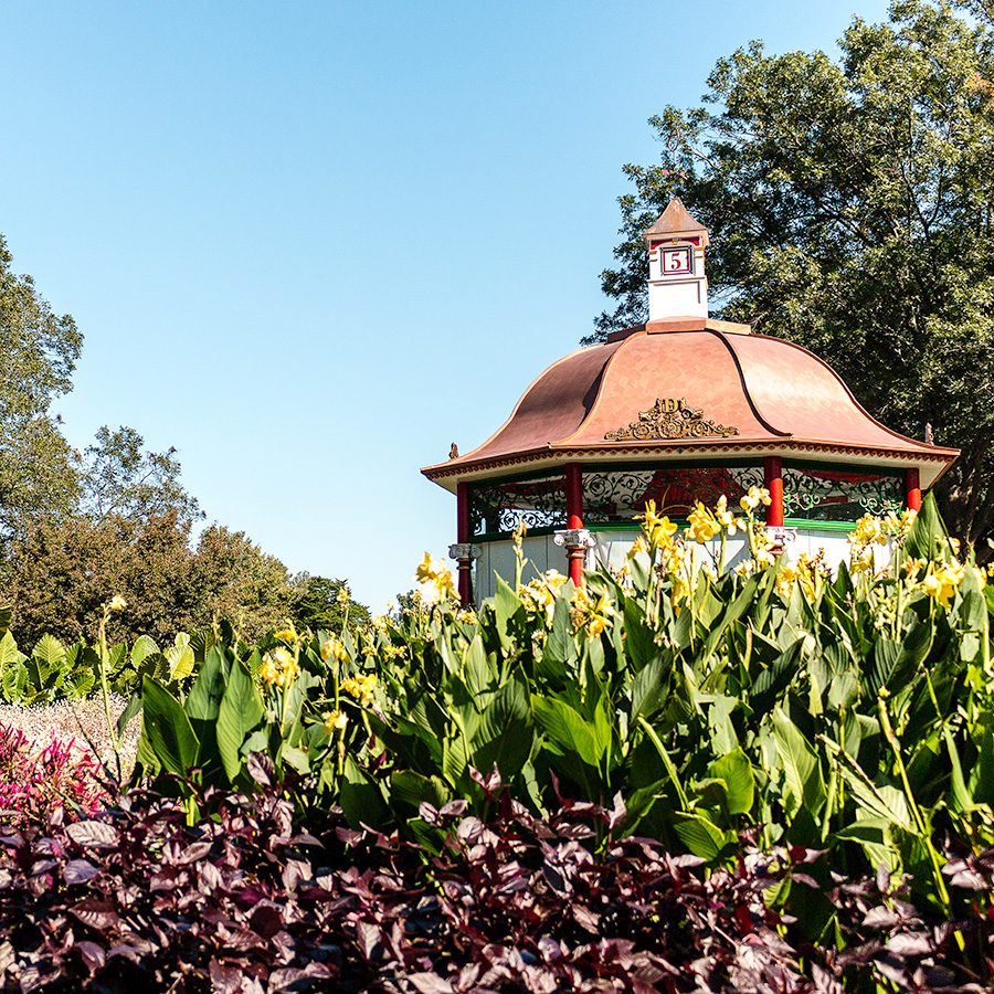 Plants and flowers surround an ornate bandstand at the Dallas Arboretum & Botanical Garden on a clear blue day.