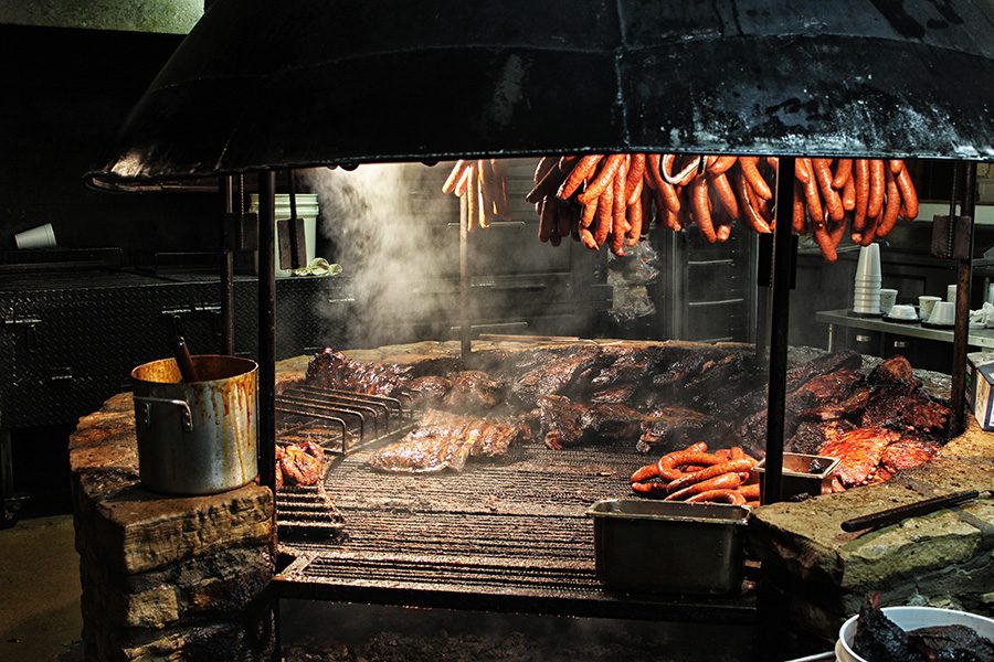 Huge cuts of beef cook on a large round covered stone barbecue pit, while sausages hang from the covering. 