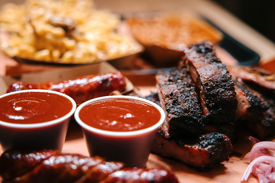A close-up of a tray of food, including slices of beef, sausages, and cups of red sauce. 