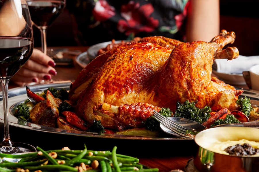 A close up of a whole roasted turkey with vegetables and a woman holding a glass of red wine at the dining table for Thanksgiving meal at Henry, Rosewood HK 