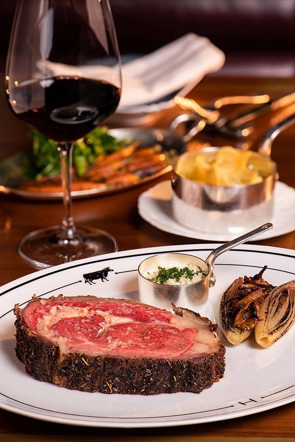 A closeup of a grilled steak on a plate with vegetables, sauce, pudding and red wine on a dining table served at Henry in Rosewood, Hong Kong 
