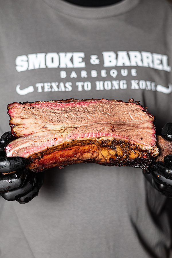 A close up of a person wearing Smoke & Barrel T-shirt holding a large smoked barbeque pork rib.