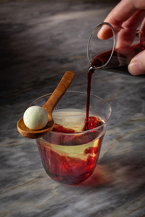 A hand pouring a red liquid into a glass of clear yellow liquid at ARGO, Hong Kong. A wooden spoon with a white ball rests on the rim of the glass. 