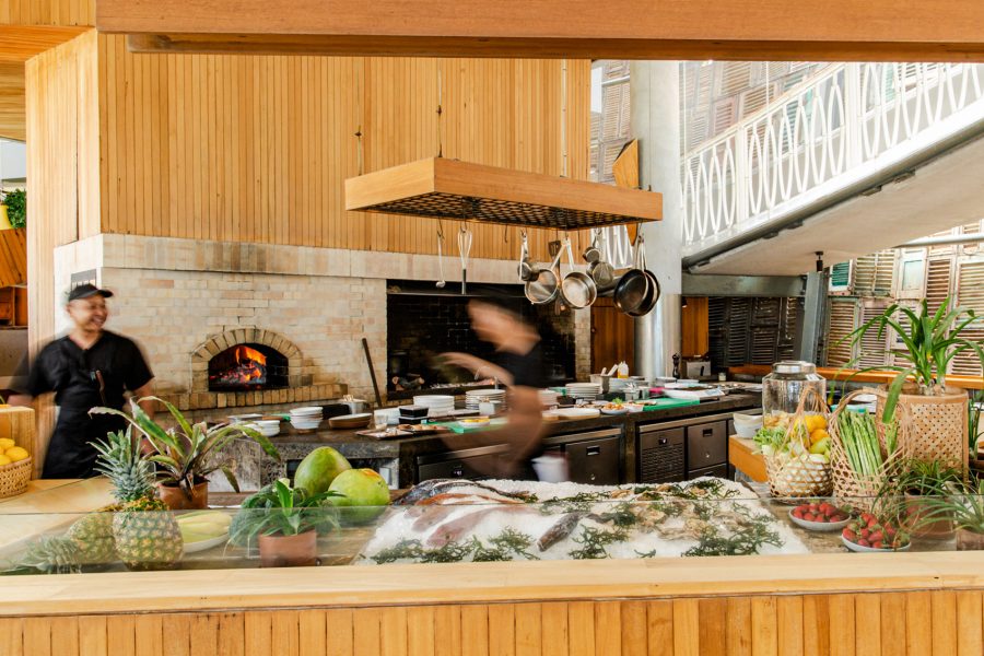 The open kitchen of Ijen at Desa Potato Head in Bali in the daytime. A tabletop is covered in fruit and fresh fish on ice. Two staff members are in the kitchen. 