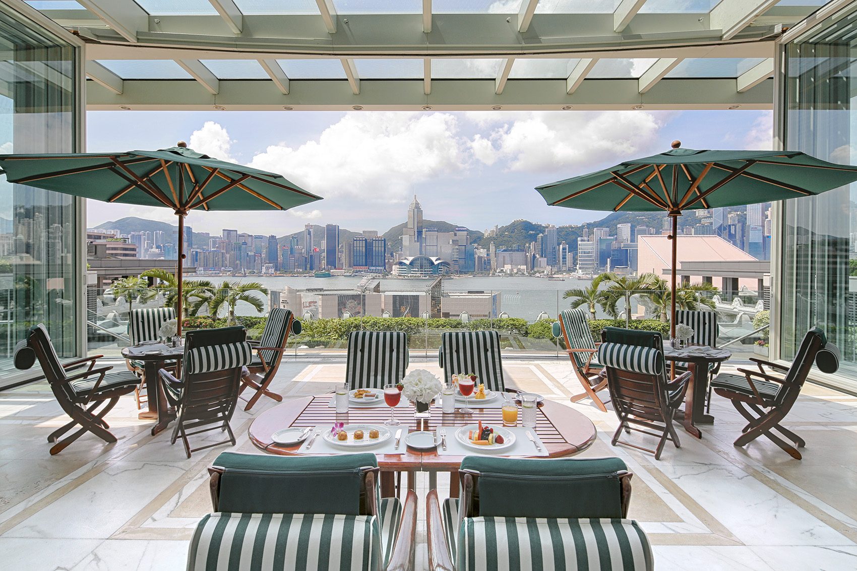 Lounge chairs and sun umbrellas in an open air lounge dining area with a view of Victoria Harbour and Hong Kong’s cityscape from The Peninsula Hong Kong 