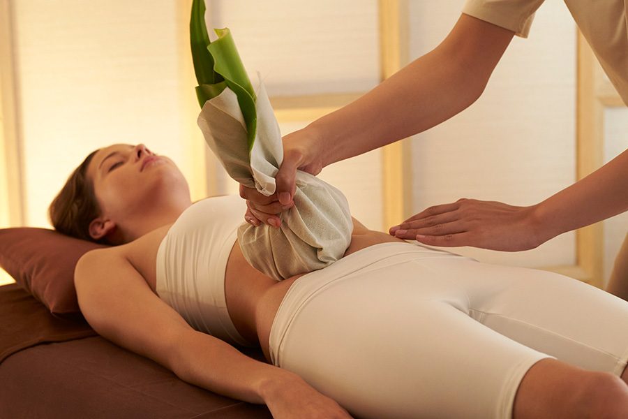 A woman wearing white lying on a massage table while a masseuse gently applies traditional salt pot therapy on the abdomen.