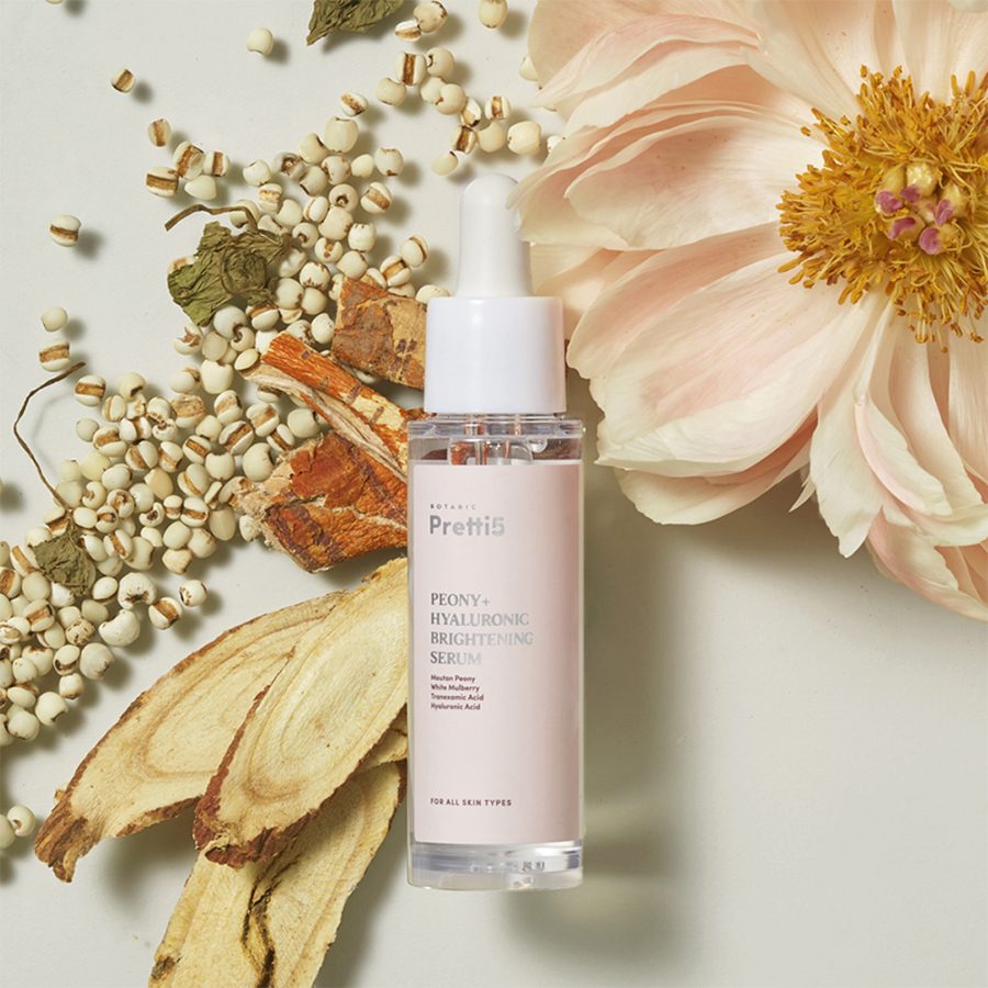 A bottle of Pretti5 Peony + Hyaluronic brightening serum with flowers and botanical ingredients in the background 