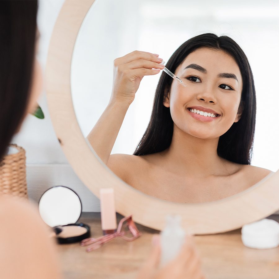 Asian woman smiling and applying serum on her face in front of a mirror 