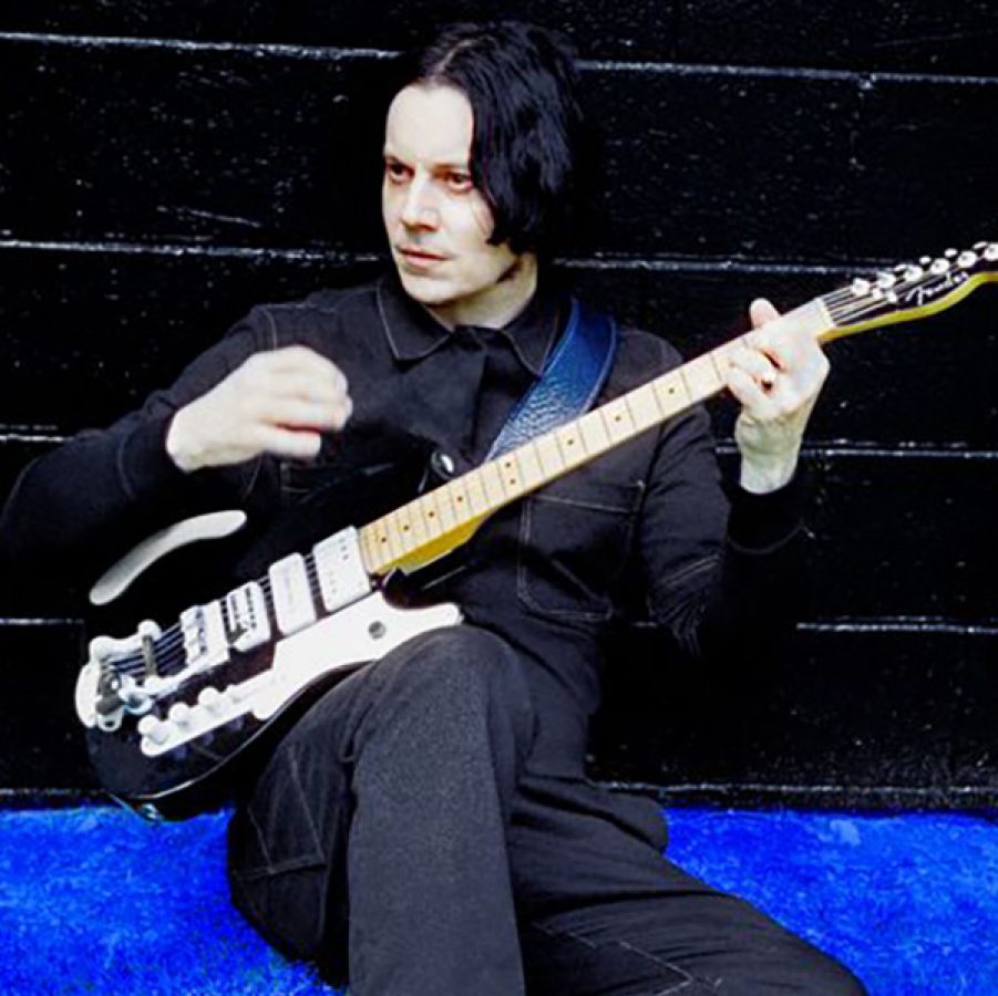 Jack White is seated against a black brick wall dressed in black and holding a black guitar.