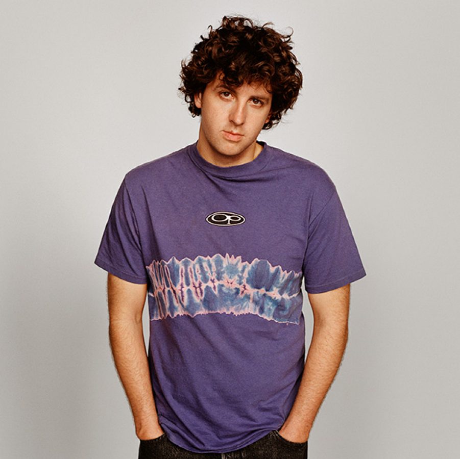 British DJ Jamie xx is wearing a purple top and black trousers against a grey wall.
