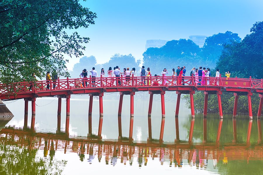 People walking across the red Huc Bridge over Hanoi’s Hoan Kiem Lake in the daytime. 