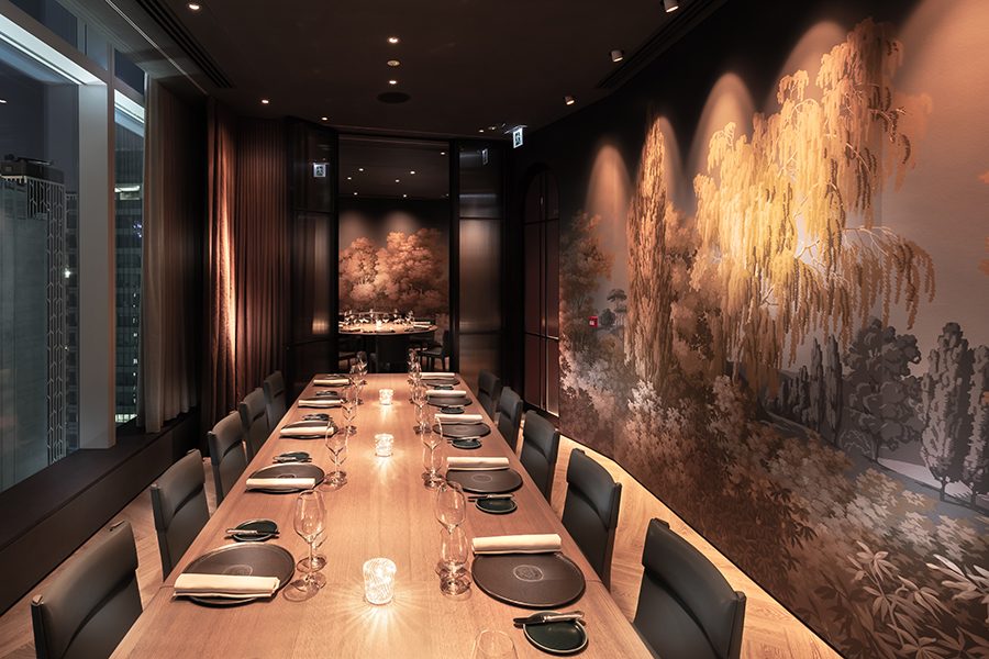 A private dining room with a long wooden dining table set with mats and napkins. One wall features a mural; another has a large window showing the city at night.