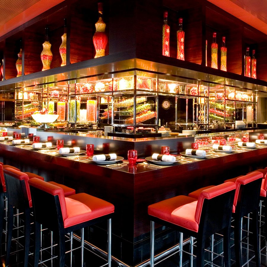 Inside a warmly lit room, a long red bar counter is lined with red bar stools and laid with plates and red glasses.