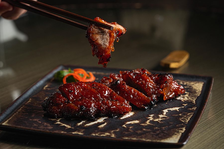 A pair of chopsticks lift a piece of char siu from a plate.