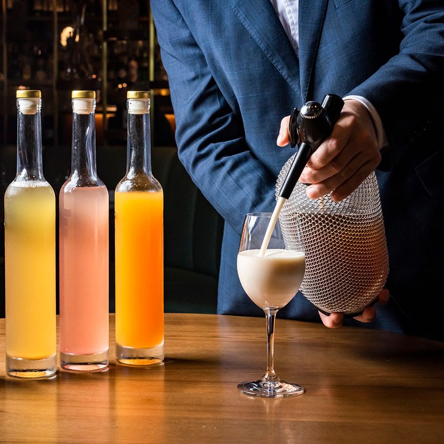 Bottles of colourful, non-alcoholic drinks are lined on a table, while a server pours a drink into a wine glass.