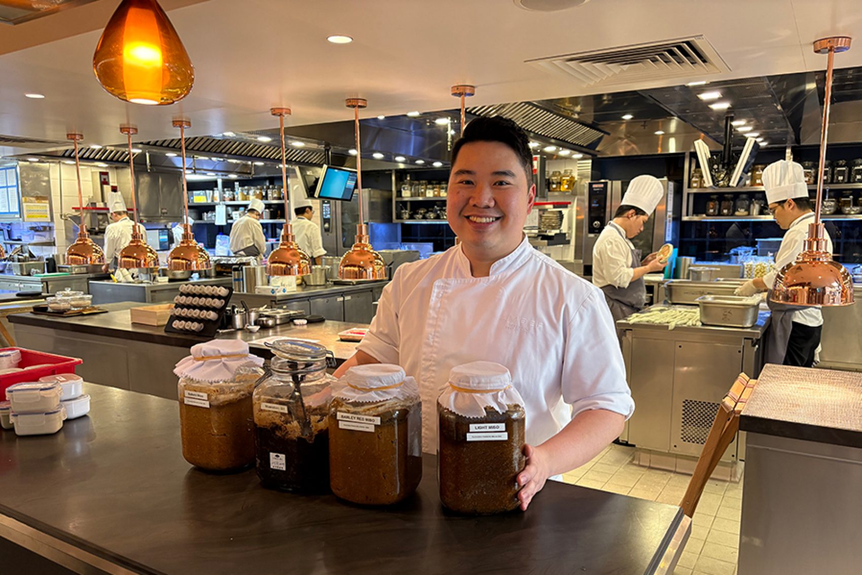 Terry Ho stands behind a counter in a restaurant kitchen. On the counter are several large jars of miso. 