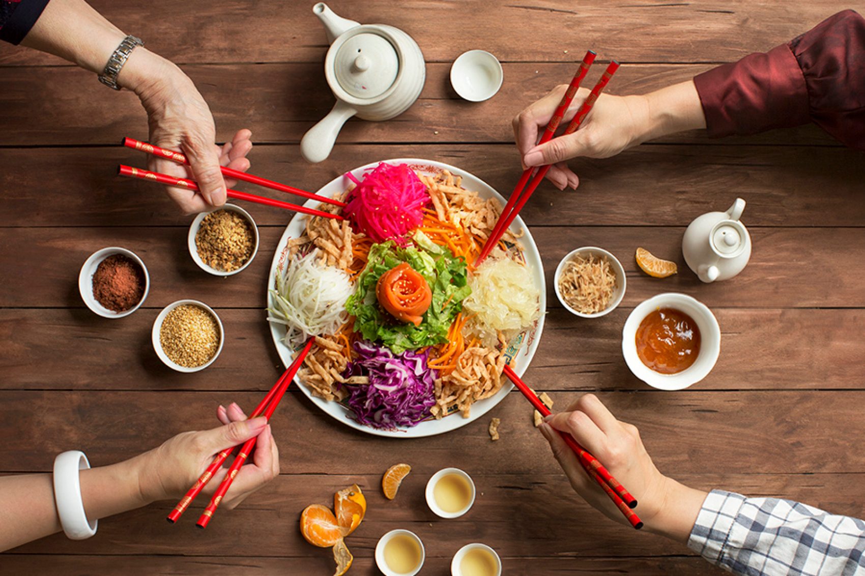 Four hands holding red chopsticks over a plate of lo hei. There are also teapots, teacups, condiment bowls and mandarin slices on the table.