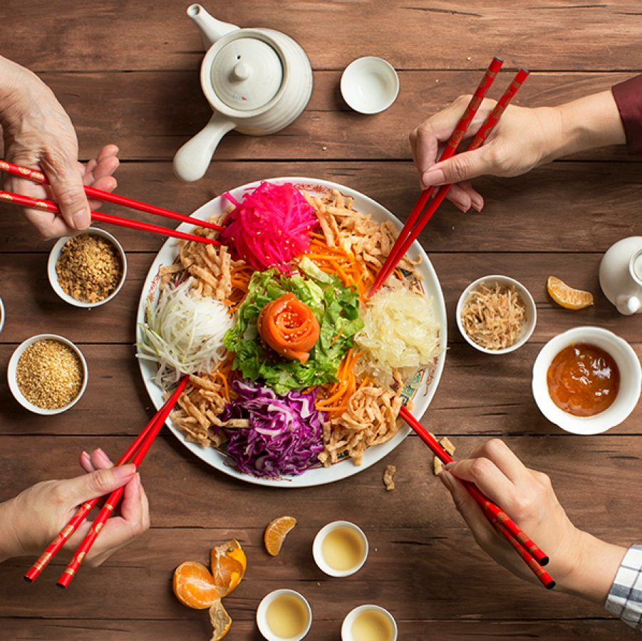 Four hands holding red chopsticks over a plate of lo hei. There are also teapots, teacups, condiment bowls and mandarin slices on the table.