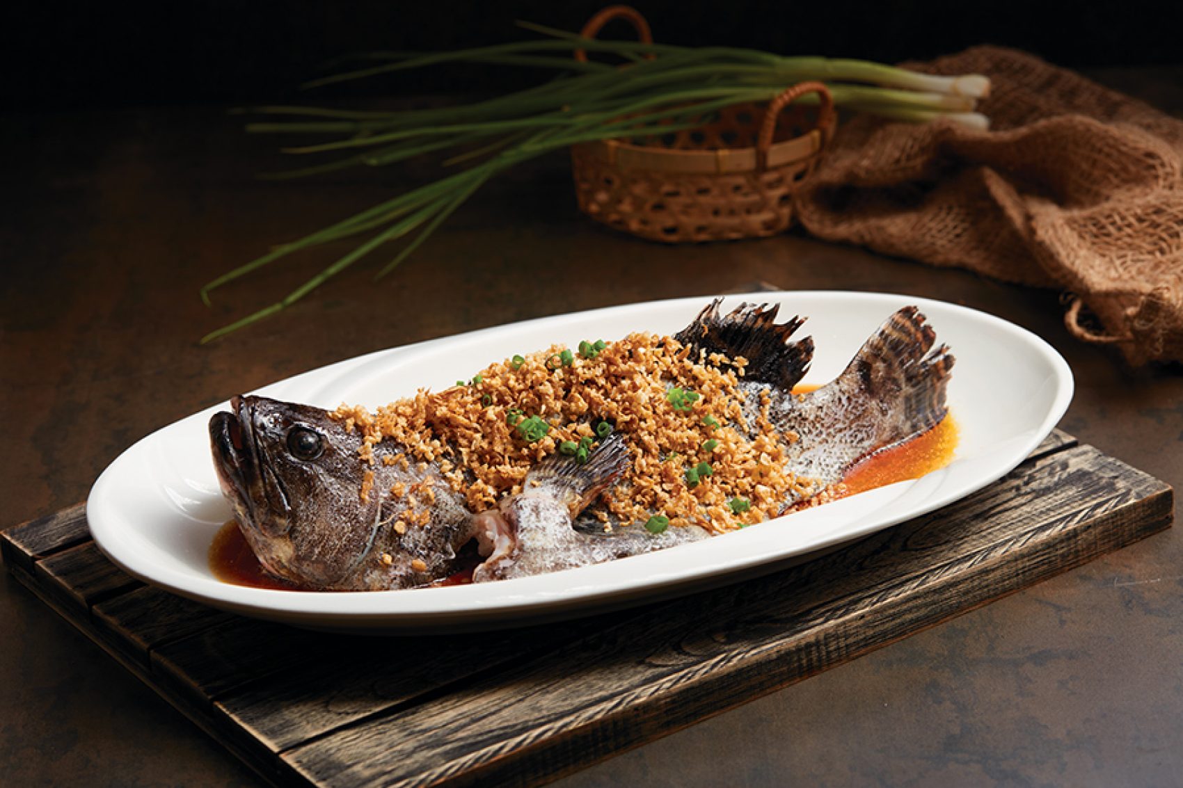 Fish on a plate with spring onions and a woven basket behind it. 