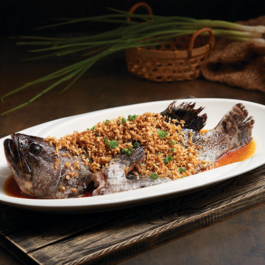 Fish on a plate with spring onions and a woven basket behind it. 