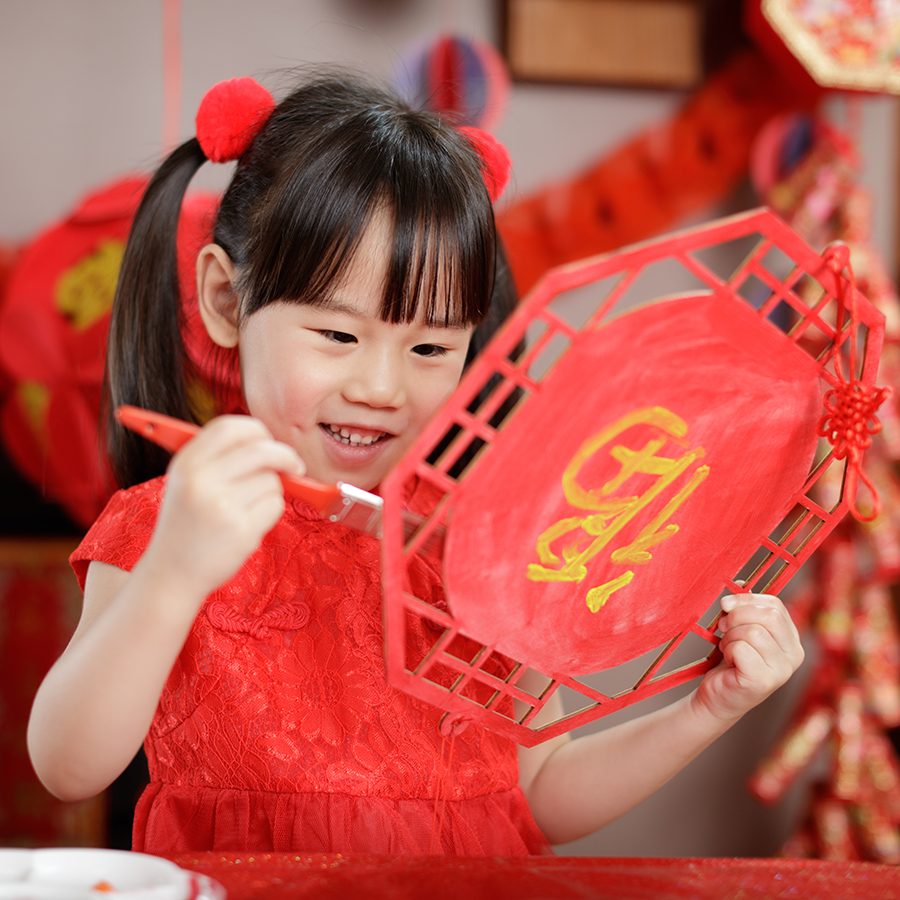 A young girl dressed in red is painting a red Chinese New Year decoration. There are many other red Chinese New Year decorations hanging behind her.