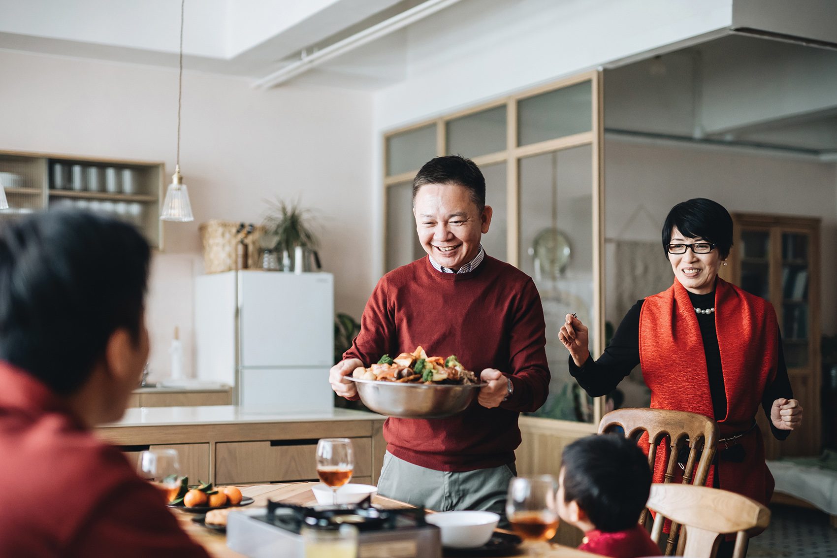 Man carrying over a bowl of food to family gathered around the dining table all dressed in red clothing.