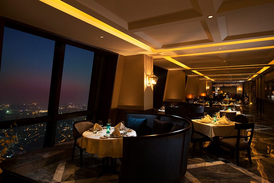 An interior view of the dining room of Italian Bridge, featuring a round table by the floor-to-ceiling window and a view of the city at night at Hilton Zhongshan Downtown.