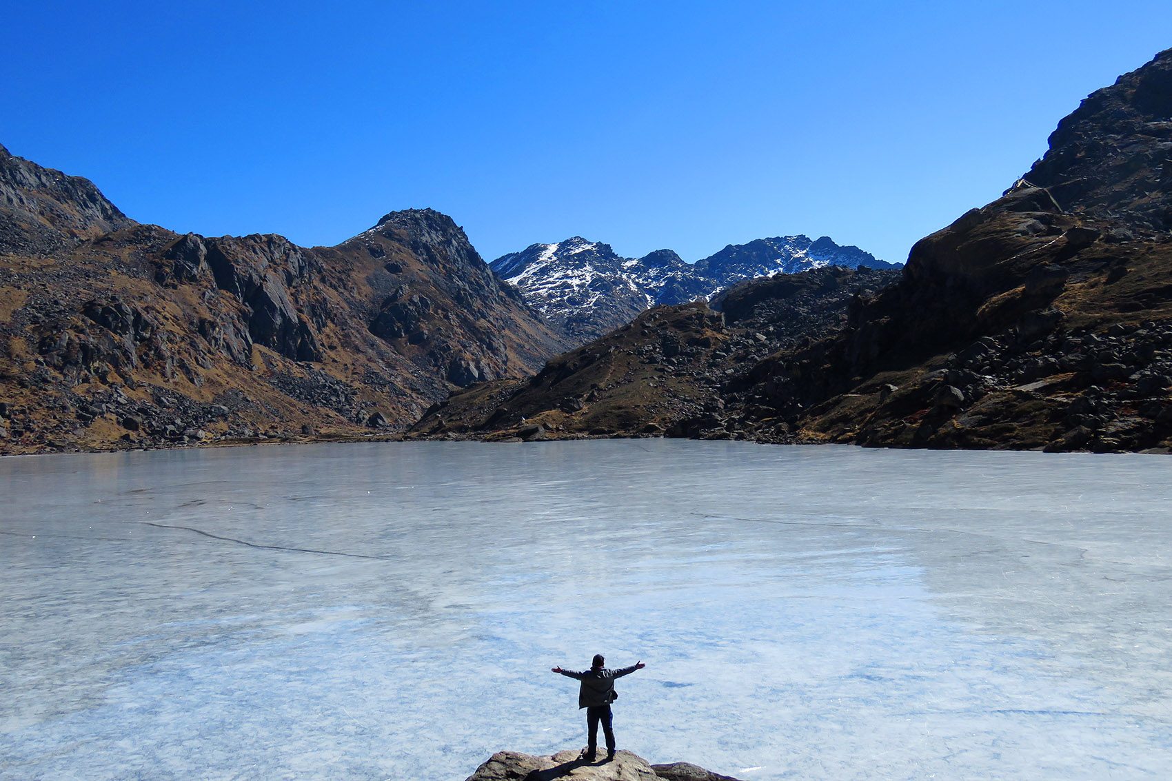 A person standing on a rock with their arms outstretched, facing Gosaikunda Lake and mountain ranges in Langtang National Park. 