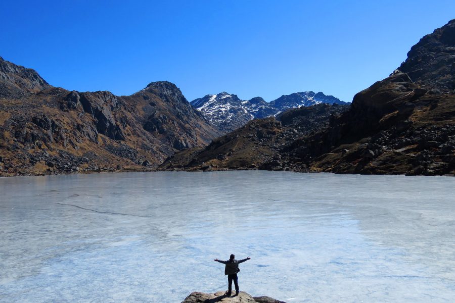 A person standing on a rock with their arms outstretched, facing Gosaikunda Lake and mountain ranges in Langtang National Park. 