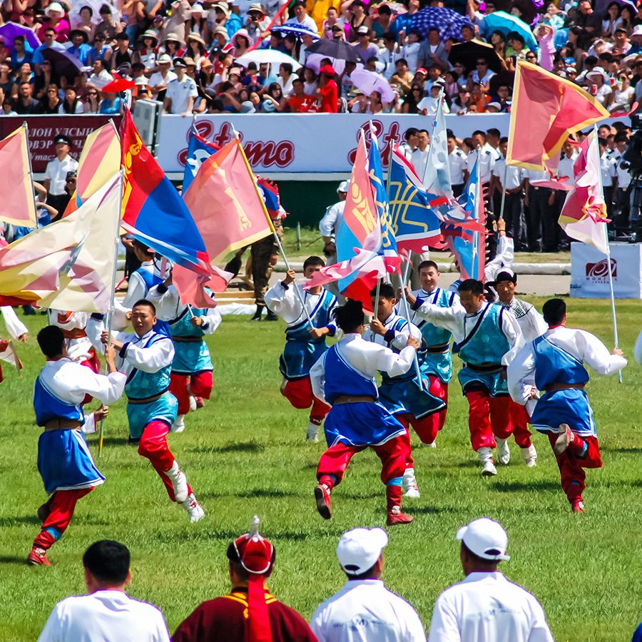 A group of men dressed in blue, red and white, running on a field while holding flags at the Naadam Festival. 