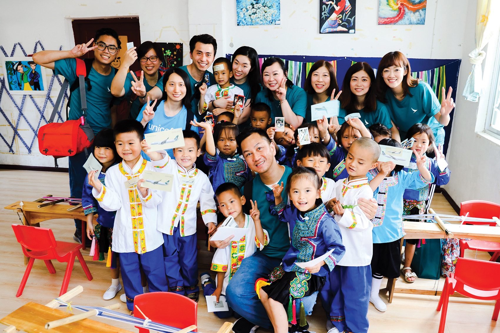 A group of happy children and Unicef Hong Kong staff smiling together and holding up a Cathay Pacific card in a classroom.