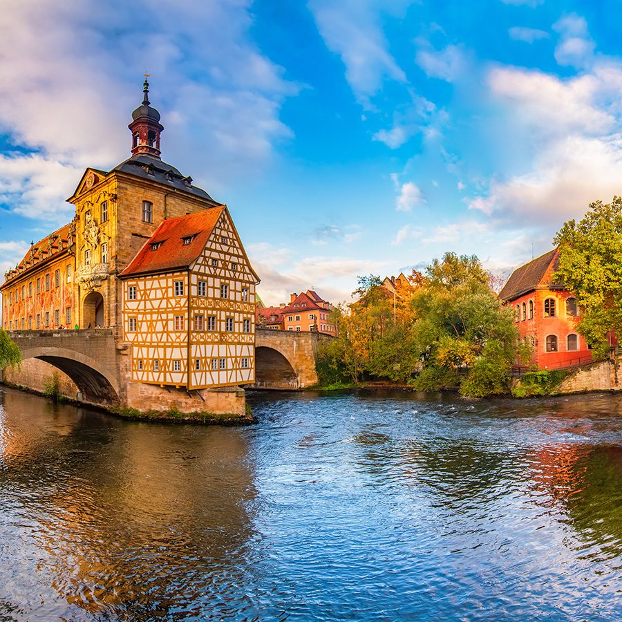 Panoramic view of Bamberg’s old town, with the Altes Rathaus, Regnitz River, colourful timber houses and foliage. 