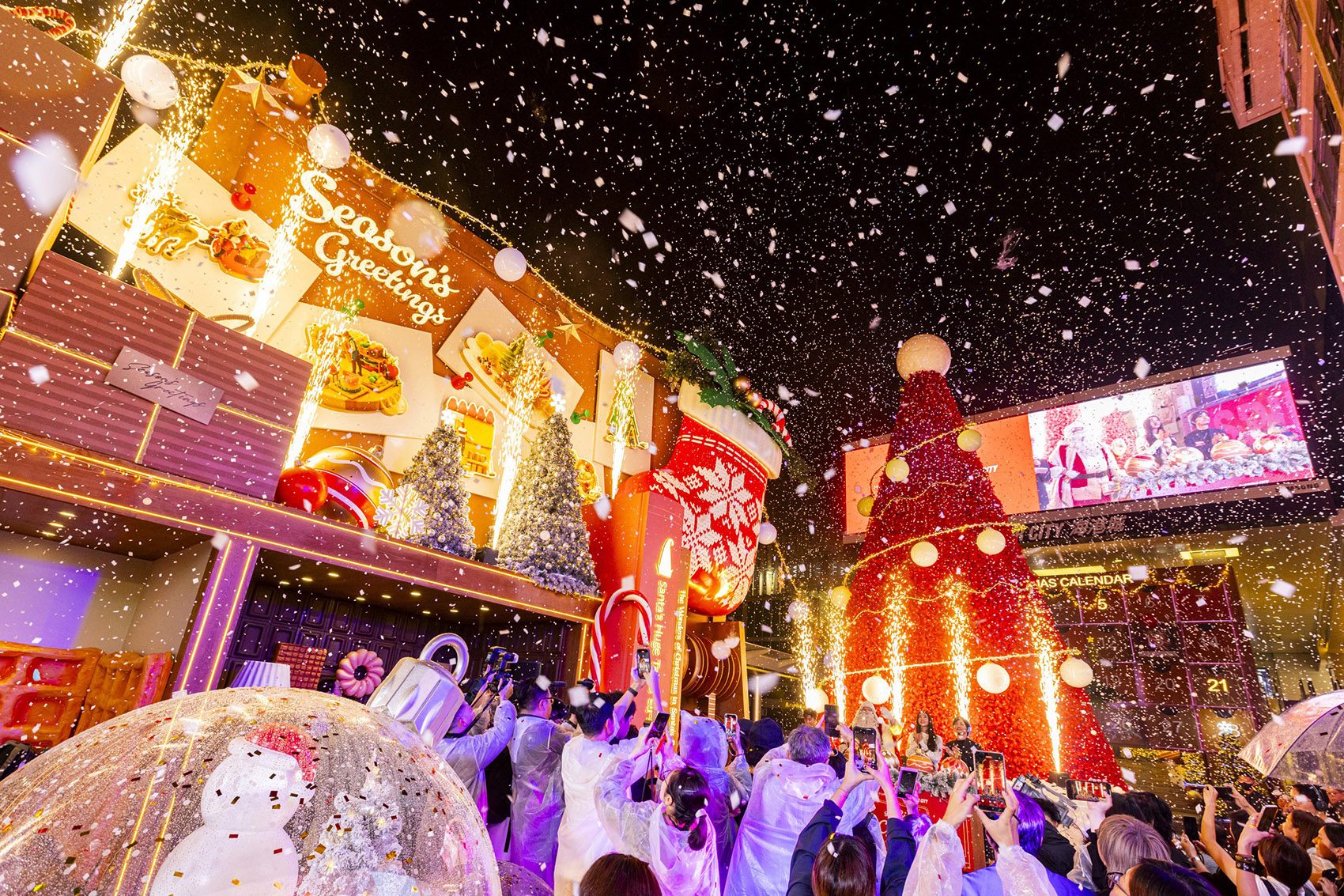 Crowds gather around an illuminated Christmas display as artificial snow falls from the sky. 