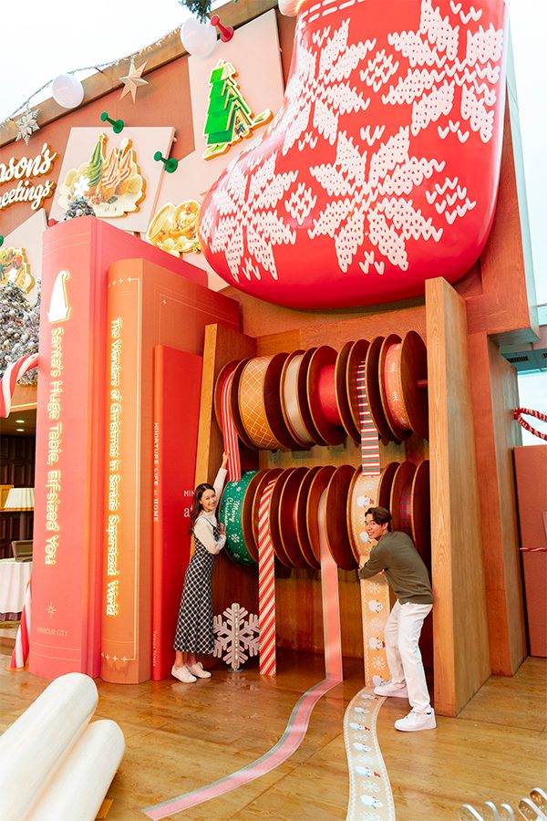 Two people pose next to a display of oversized rolls of ribbon at the Big Little Christmas installation at Harbour City.