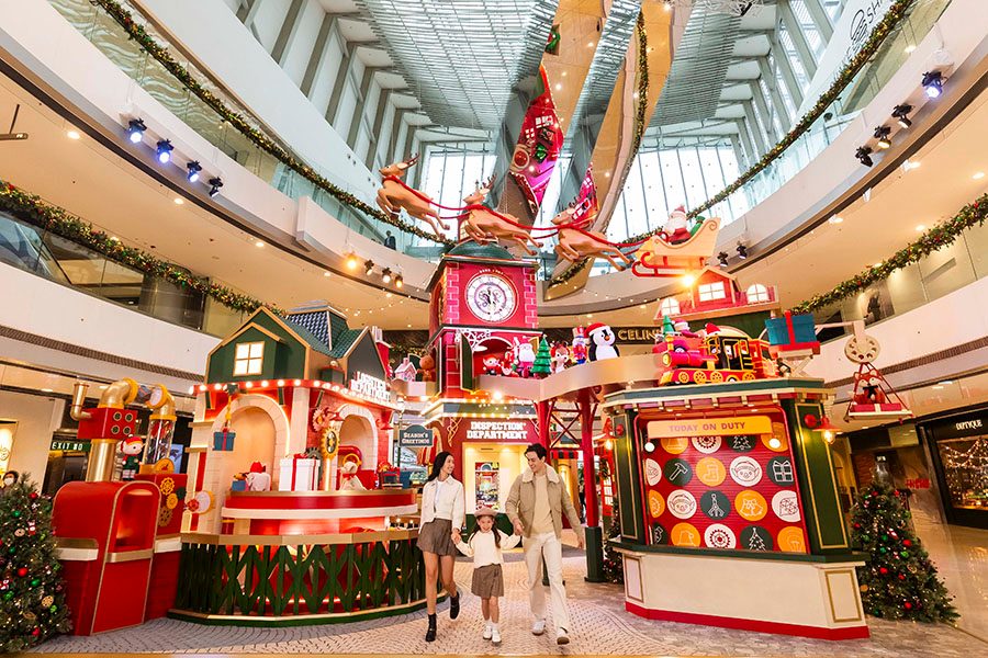 A family stands in front of the IFC’s Santa’s Magic Toy Factory display. It comprises several red and green buildings and a red clock tower, over which a model sleigh and reindeer are suspended. 