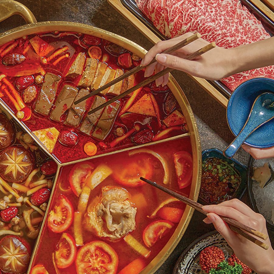 A hotpot with three compartments filled with different kinds of broth, mushrooms, tomatoes and other ingredients at Coucou Hotpot · Tea Break, Hong Kong. Plates of raw meat, shellfish and watermelon are on the table surrounding the pot. Hands holding chopsticks and a bowl can be seen near the pot.