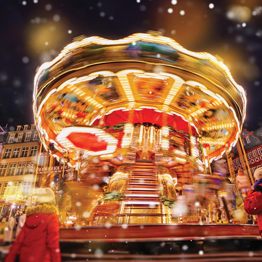 A crowd watching a moving carousel with Christmas decoration and snow falling at the Frankfurt Christmas Market at night.