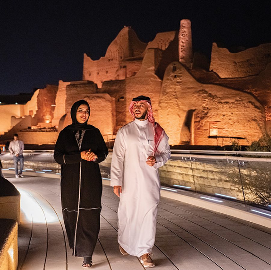 Man and woman walking along a path with illuminated architecture behind them in Riyadh.