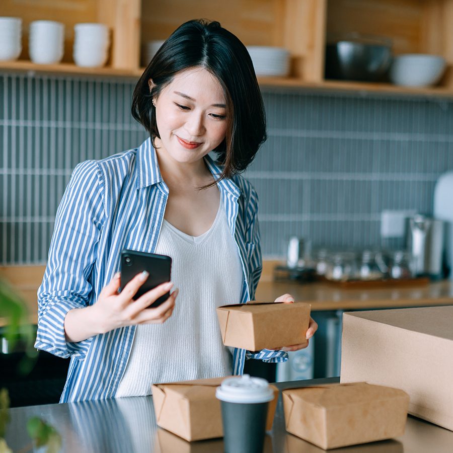 A woman in the kitchen with takeaway boxes and a cup while holding a mobile phone and smiling