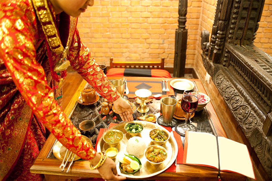 A woman in traditional Nepali dress sets down a platter of daal bhaat on a table. 