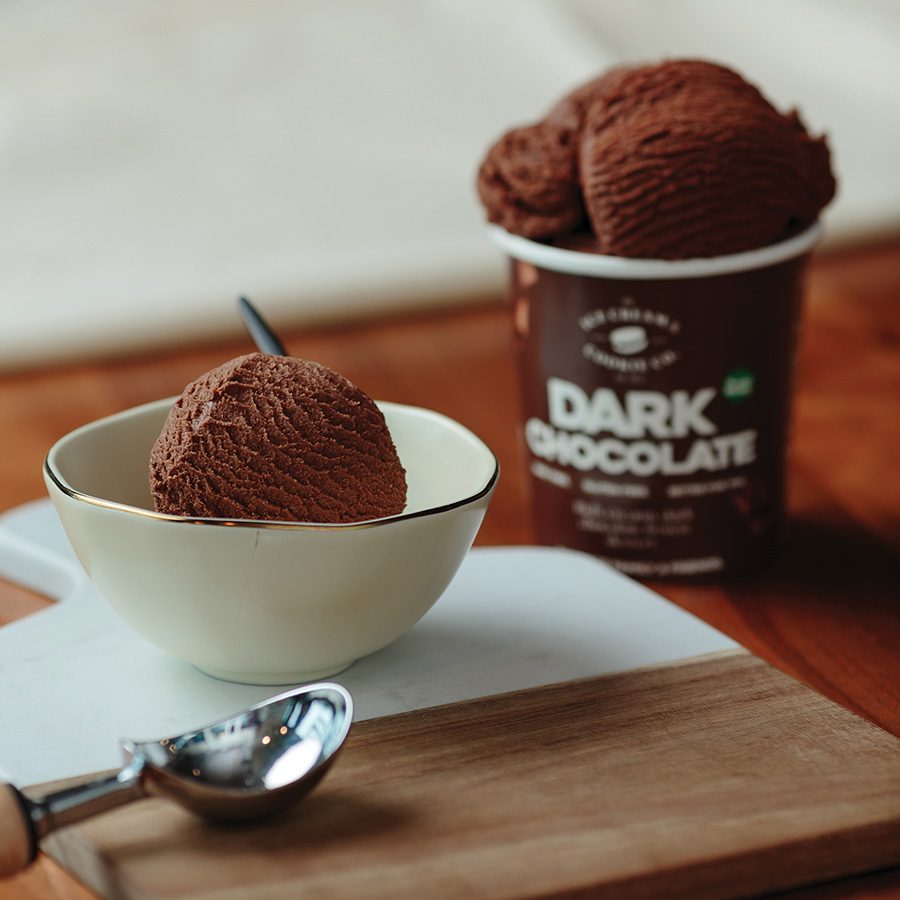 A closeup of a scoop and pint of dark chocolate ice cream from Ice Cream & Cookie Co, served on a wooden table.