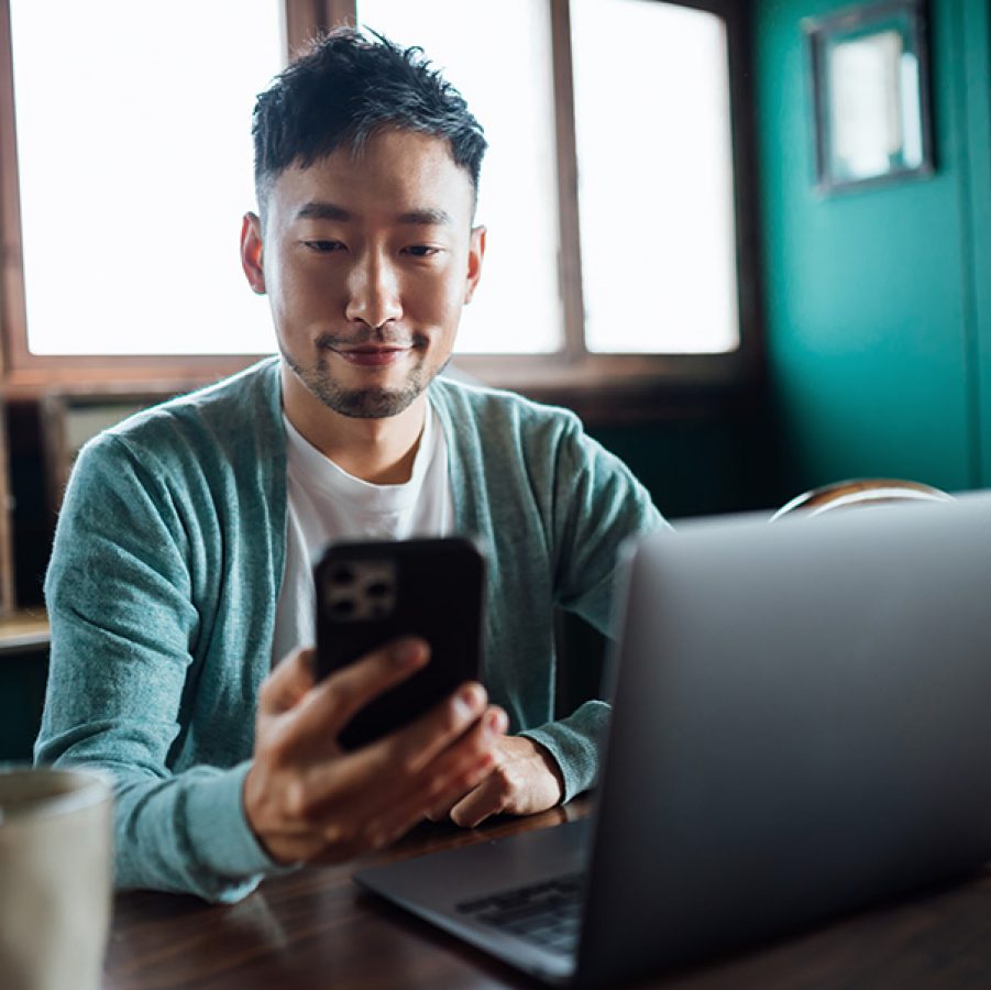 A seated man smiling as he looks at the screen of his phone with an open laptop and a cup on the table in front of him.