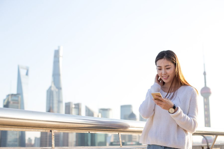 A woman smiling and looking down at her phone while outdoors, the skyline of Shanghai behind her in the distance. 