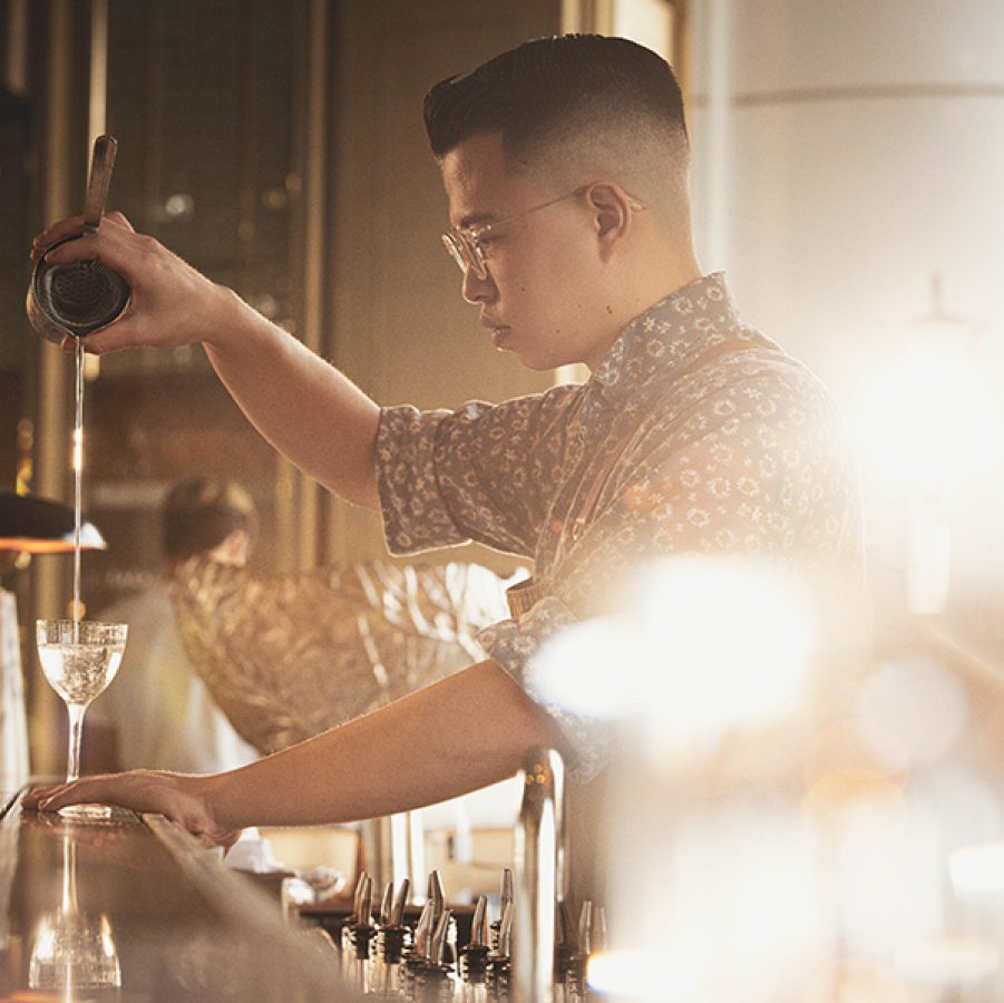 A side profile of ARGO senior bartender Evan Mak behind the bar pouring a cocktail into a glass.