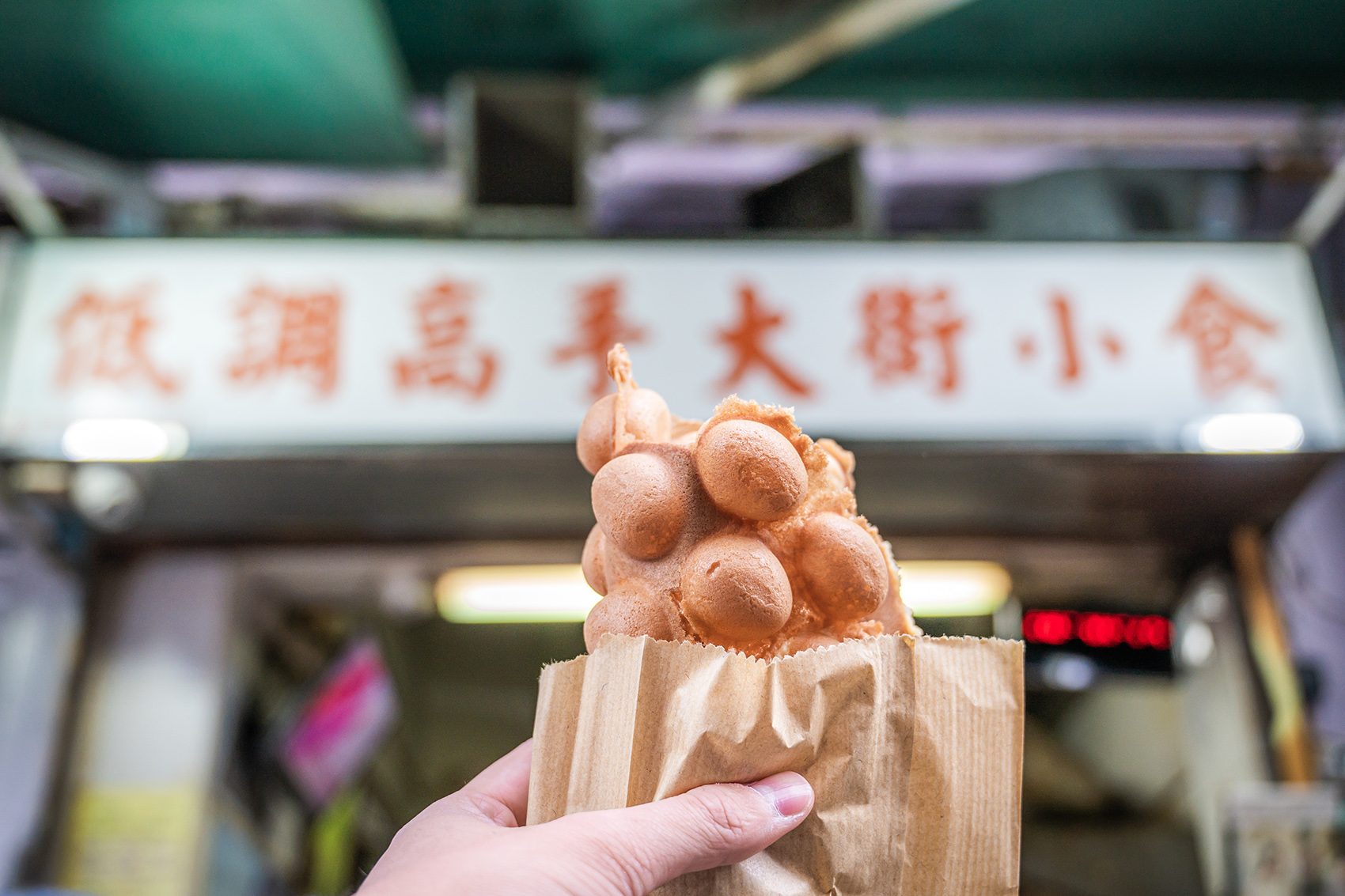 A hand holding a paper bag of egg waffles in front of Master Low-Key Food Shop.  